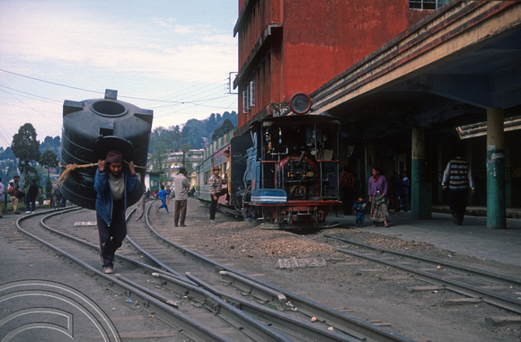 T6904. Porter carrying a eater tank past the toy train. Darjeeling. West Bengal. India. April.1998.